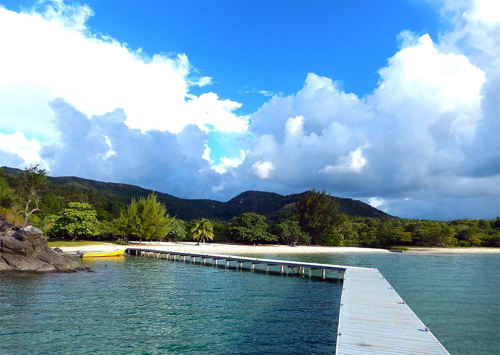 Guanaja Faqs - Things You Need To Know Before You Go (2022) 10 white jetty across blue sea to a sandy beach with bushes and trees behind and blue skies