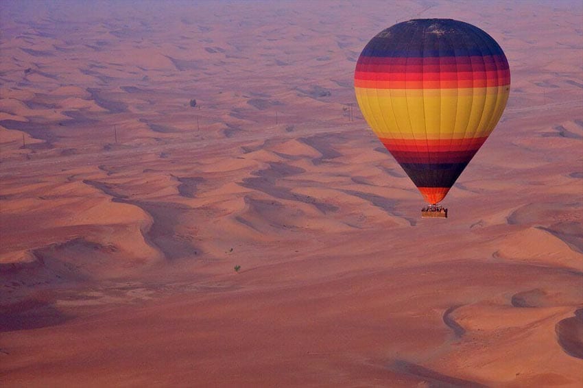 A Week In Dubai Itinerary - Best Things To Do In Dubai In 7 Days 6 Hot air balloon with bands of purple, pink red orange and yellow and basket beneath flying over red sand dunes in the desert