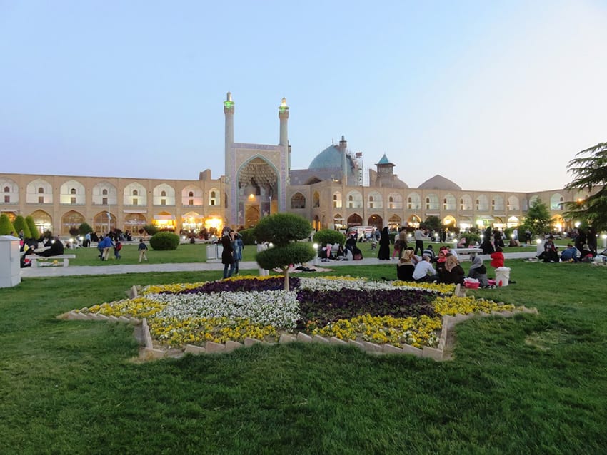 8 Amazing UNESCO Sites in The Middle East - Off the Beaten Track 4 Naqs-e Jahan Square with arched sand coloured buildings in the background with three domes on the right and a larger arched entrance in the middle. In the foreground is a grassy area with a seven sided star planted with white, purple and yellow flowers and a tree in the middle. People are sitting on the grass or walking in the grounds.