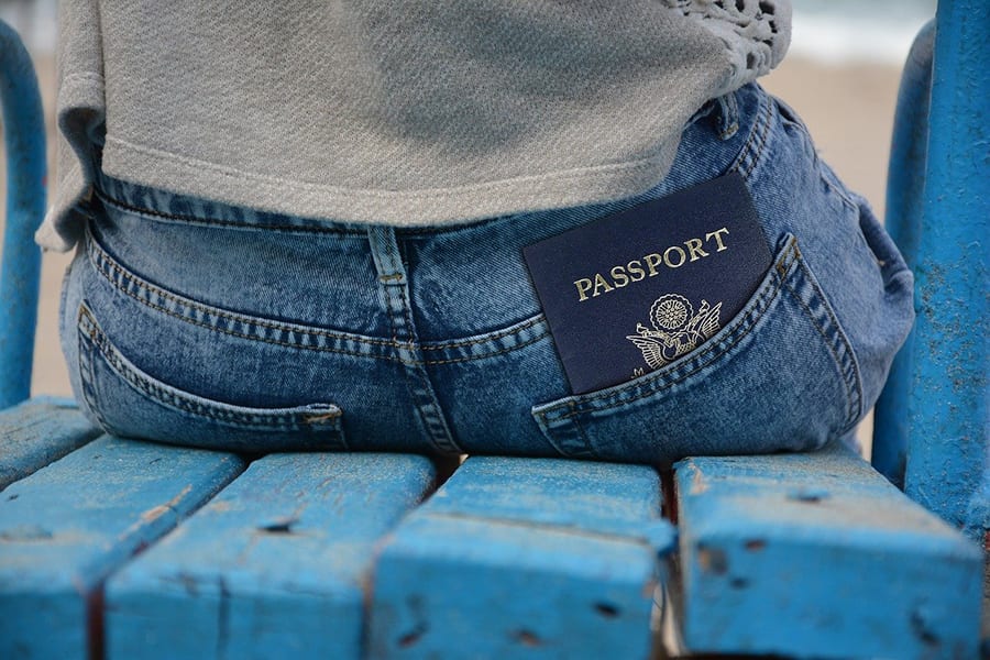 19 Awesome Solo Travel Tips For When You Travel On Your Own 8 image of the back of a woman wearing blue jeans and a grey sweatshirt sitting on a blue wooden seat with a blue passport in her jeans pocket