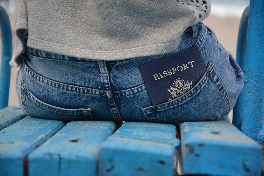 image of the back of a woman wearing blue jeans and a grey sweatshirt sitting on a blue wooden seat with a blue passport in her jeans pocket