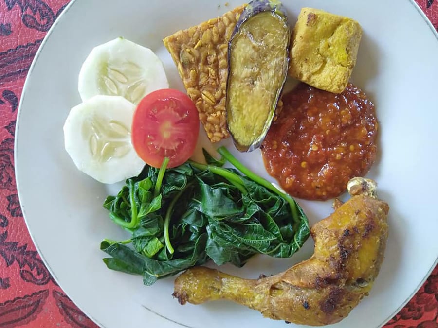 How to Cook Ayam Goreng, delicious Fried Chicken, Bali Style 3 Ayam Goreng - Balinese Fried Chicken on a white plate on red tablecloth with fried chicken leg, greens, sliced tomato and cucumber, tempe, fried aubergine, corn fritter and tomato sambal