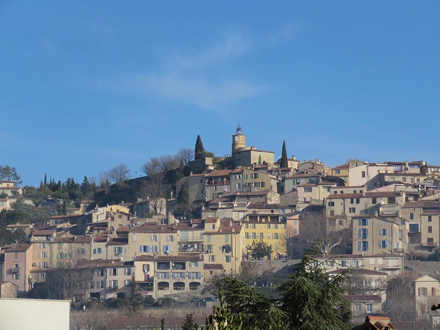 View from below of the hilltop town of Fayence, showing the terraces of cream coloured buildings with red roofs against a blue sky