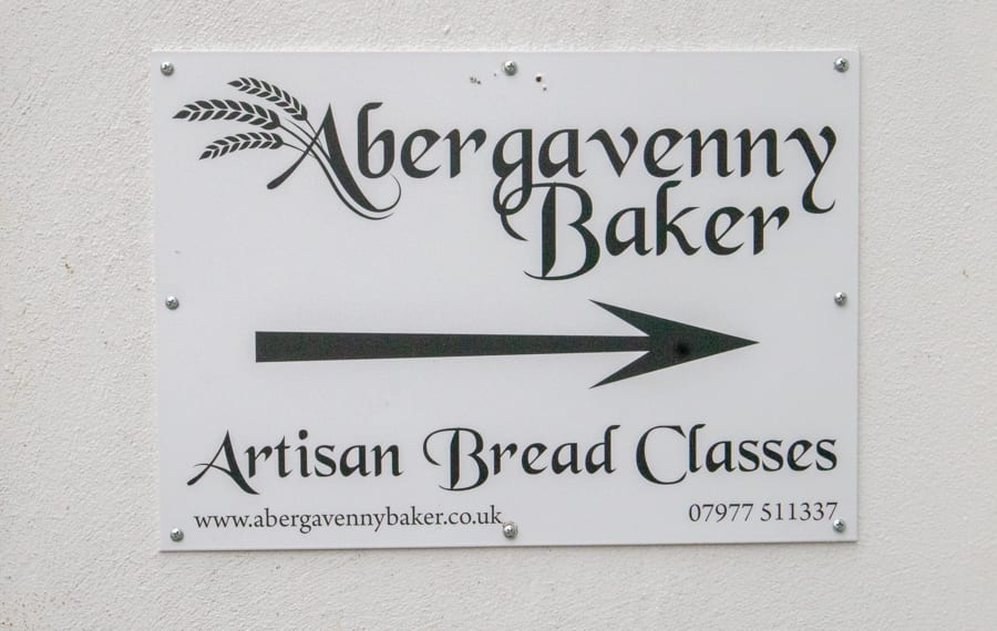 Artisan Bread Baking at the Abergavenny Baker 3 White sign on white wall with black writing saying Abergavenny Baker the A has three wheat ears coming off to the left. Below is a black arrow pointing to the right and Artisan Bread Classes below