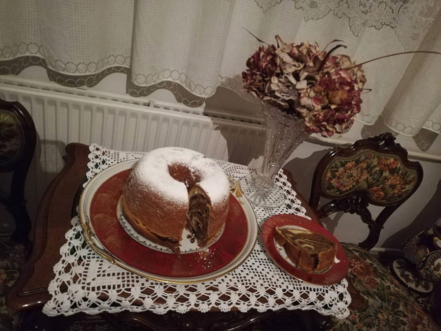 Favourite Christmas Bakes to try this year 10 round marbled potica cake from Slovenia dusted with icing sugar and sitting on a red plate. The table has a lace tablecloth and there is dried flowers and net curtains in the background