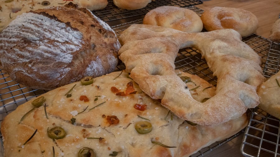 Artisan Bread Baking at the Abergavenny Baker 2 Brown loaf, fougasse bread with holes in it and focaccia with loives, tomatoes, salt and rosemary sitting on wire cooling racks