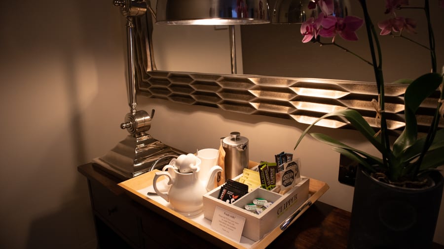 Tray with white teapot, silver cafetiere and a selection of tea sitting under a silver framed mirror and next to a pink orchid