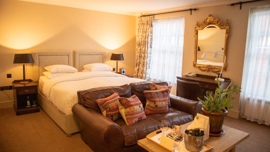 double bed with white sheets in front is a brown leather sofa with cushions in autumnal colours.  There is a gold rimmed mirror on the wall between two windows. Underneath is a desk. In front of the sofa is a table with plant and tray.