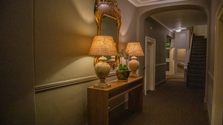 Two lamps on a side table under an orate mirror against a green painted wall with a staircase in the background