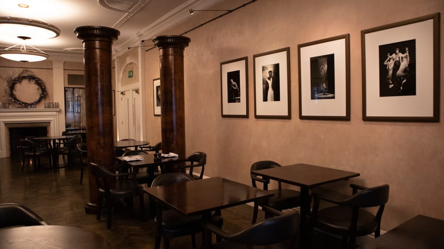 Room with pale cream walls and four framed black and white photos. There are two pillars and brown tables and chairs. In the background on the left is a fireplace with a round wreath above it.