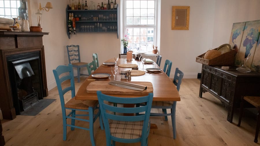Artisan Bread Baking at the Abergavenny Baker 11 wooden table with blue painted chairs . There is a fireplace on the left and a low unit with a cane dome on top.