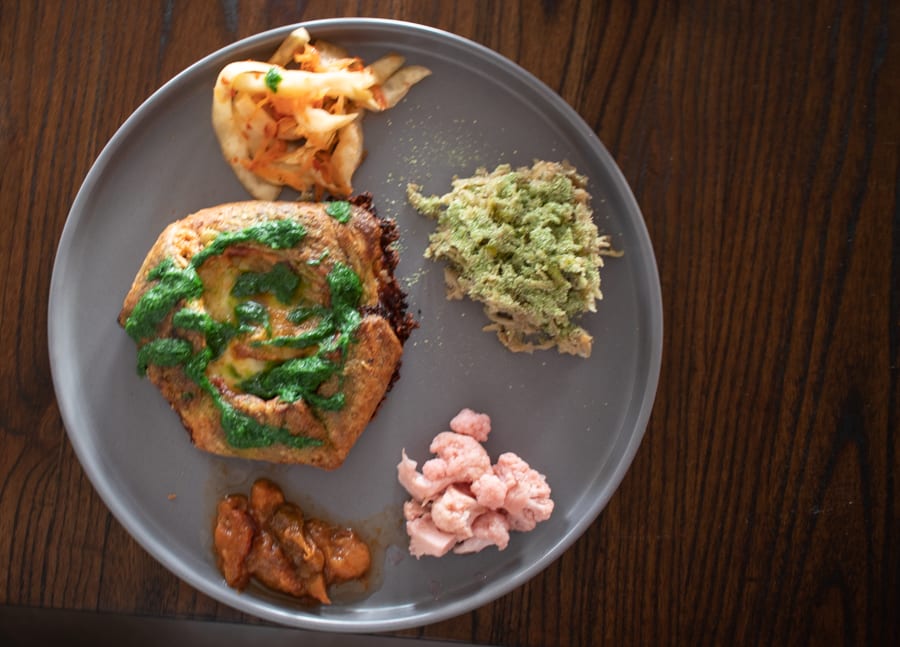 Versa - Responsibly Sourced Dining On The Isle Of Man 10 plate with potato galette covered in green wild garlic sauce surrounded by cabbage and carrot kimchi, pickled celeriac, pink pickled cauliflower and damson pickle