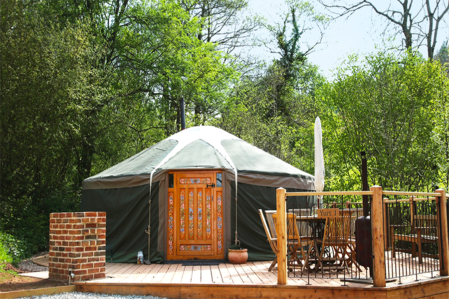 Yurt with traditional door sitting on a wooden patio with table, chairs and umbrella with trees in the background