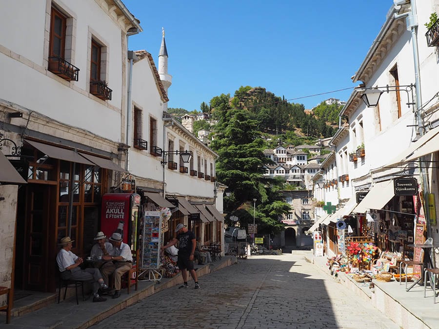 35+ Amazing Hidden Gems In Europe You Must Visit 31 street in Gjirokastra with white buildings either side and souvenirs on the pavement under awnings