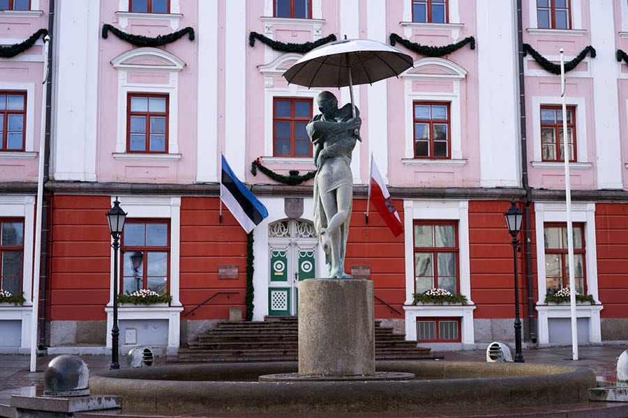 35+ Amazing Hidden Gems In Europe You Must Visit 29 Tartu Town Hall Square with pink and red building behind a fountain with a sculpture of a couple wrapped around each other kissing with the man holding an umbrella above them