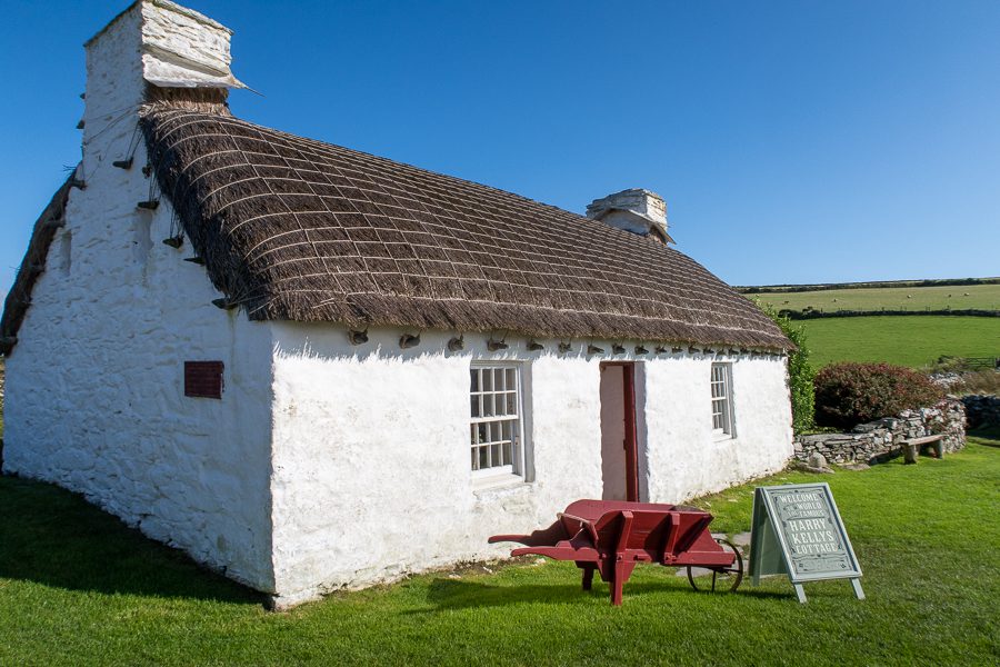 Typical Manx white stone cottage with thatched roof. There is a red painted wheelbarrow outside the door and a green painted sign