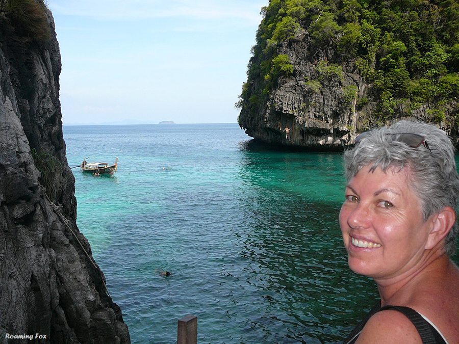 Celebrating 16 Awesome Mature Bloggers In 2021 To Inspire You 7 Alma from Roaming Fox on PhiPhi Island, Thailand smiling at the camera with turquoise sea and green covered rocks behind her