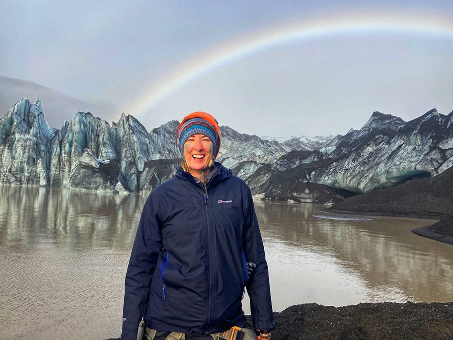 Celebrating 16 Awesome Mature Bloggers In 2021 To Inspire You 8 Sue from SueWhereWhyWhat in Iceland wearing a blue jacket and an orange and turquoise woollen hat with mud on her cheeks,with icy cliffs behind her and a rainbow in the sky
