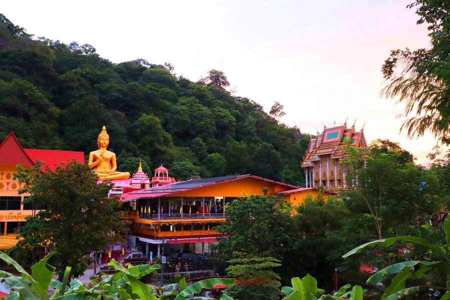 25 Amazing Destinations For Winter Sun Around The World 5 a large golden buddha next to brightly painted buildings in pinks, purples oranges and reds in amongst green trees