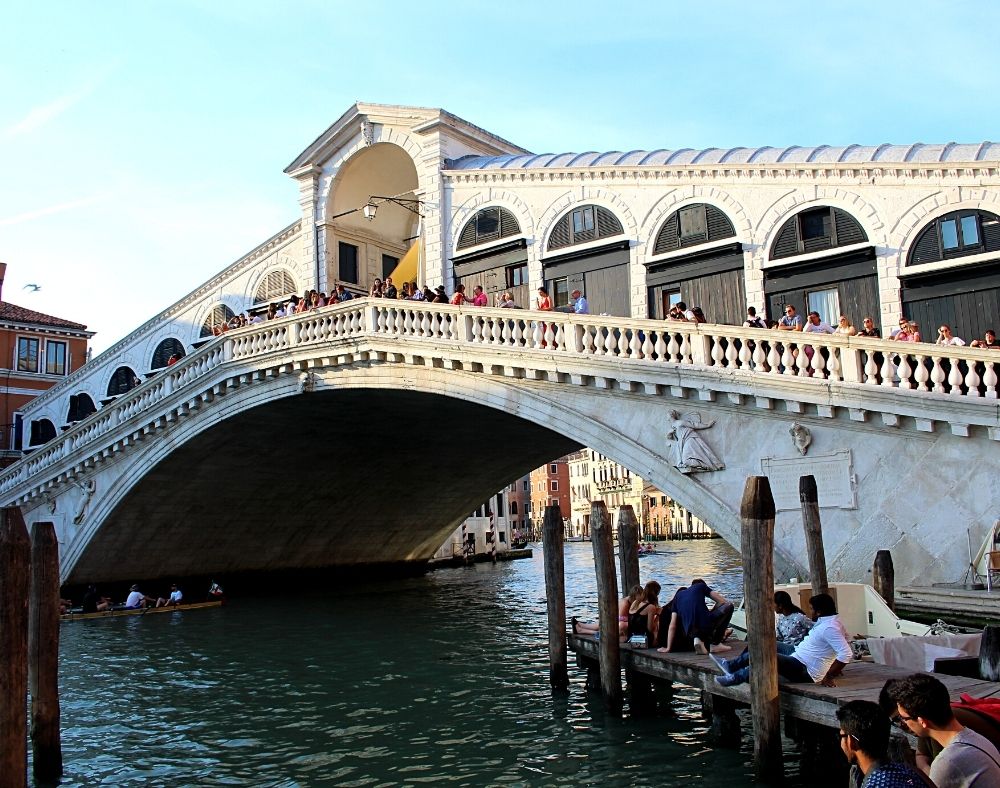 Stunning UNESCO Sites in Italy to discover in 2023 6 The white Rialton Bridge in Venice with arched doors and people on the bridge as a gondola goes underneath