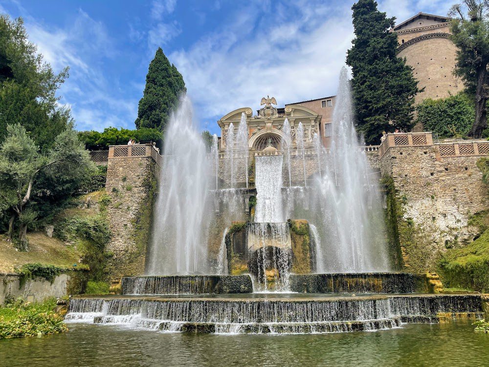Stunning UNESCO Sites in Italy to discover in 2023 18 Fountain at Villa d'Este with top level fountains, a waterfall and two main fountains either side flowing into pools below. Behind are cream coloured buildings and green cypress trees