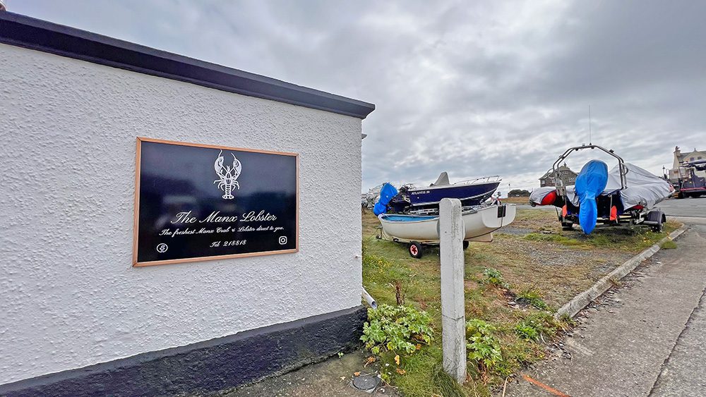 20+ Of The Best Things To Do In Port St Mary, Isle Of Man (2023) 20 White building with a black sign saying The Manx Lobster in white and a drawing of a lobster. two boats to the right with their engines covered up.