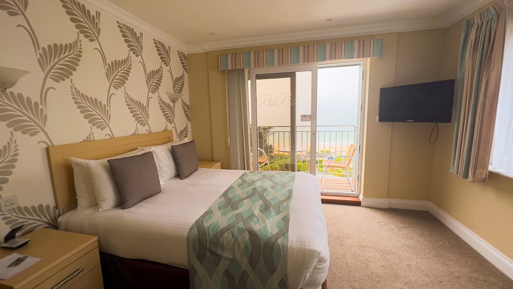 Visiting The Beautiful Biarritz Hotel In Jersey, Channel Islands (2023) 3 The bedroom at the Biarritz Hotel in Jersey with beige and cream leaf wall paper, pale yellow walls and king size bed with white linen, chocolate coloured cushions and blue, brown and white blanket over the bed. There is a TV screen on the wall in the corner and open patio windows showing a chair and table and the sea view beyond