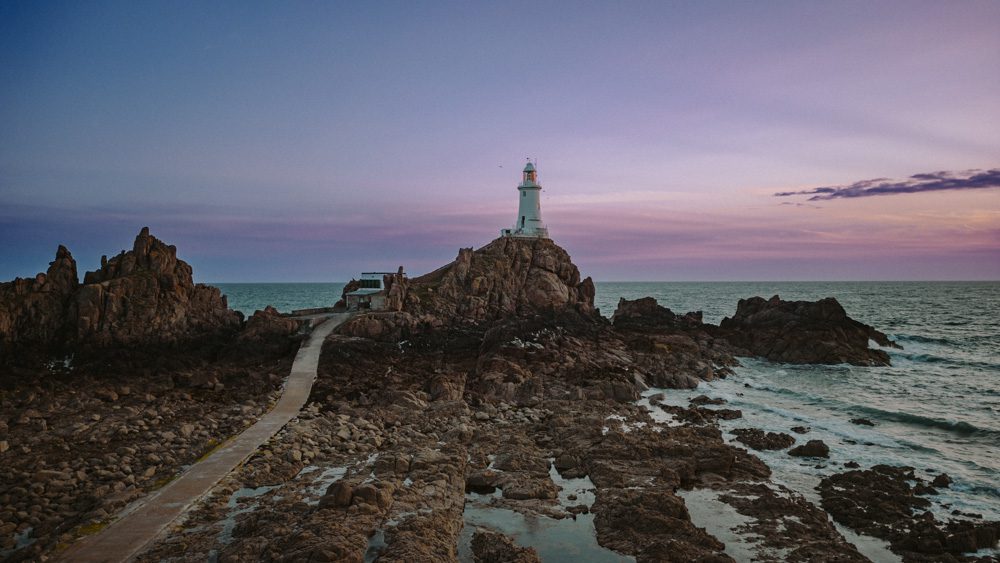 La Corbiere Lighhtouse in Jersey in the Channel Islands at Dusk with a purple and pink sky behind and the lighthouse on top of rocks. There is a cement walkway to the left and the incoming tide to the right. In the background, the sea continues to the horizon