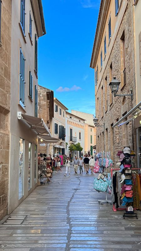27 Of The Best Things To Do In Alcudia, Mallorca (2023) 2 Narrow street in Alcudia old town with shops to the sides selling bags, hats etc