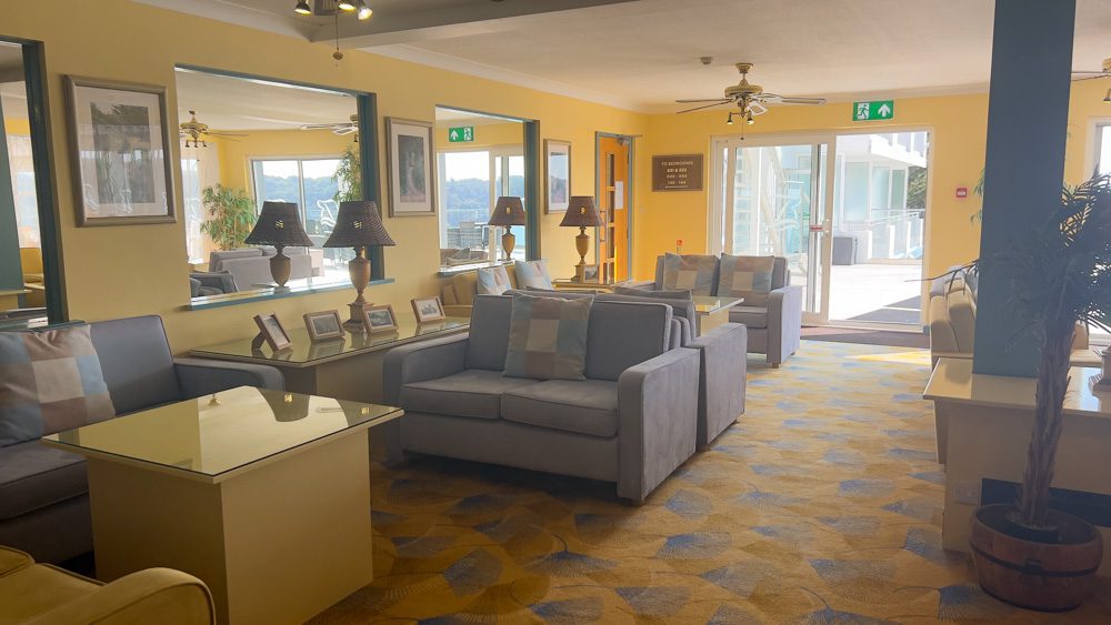 Visiting The Beautiful Biarritz Hotel In Jersey, Channel Islands (2023) 8 One of the lounges in the Biarritz Hotel with blue-grey couches, blue and yellow carpet and yellow walls with an open sliding door to the terrace