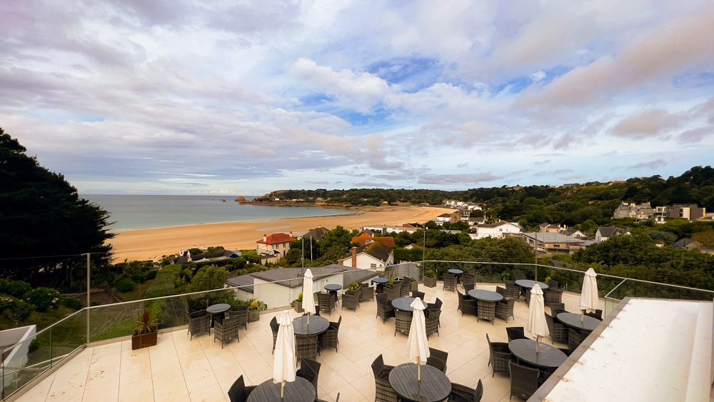 Visiting The Beautiful Biarritz Hotel In Jersey, Channel Islands (2023) 10 black wicker chairs around matching tables with white sun umbrellas on the terrace at the Biarritz Hotel overlooking St Brelade's Bay