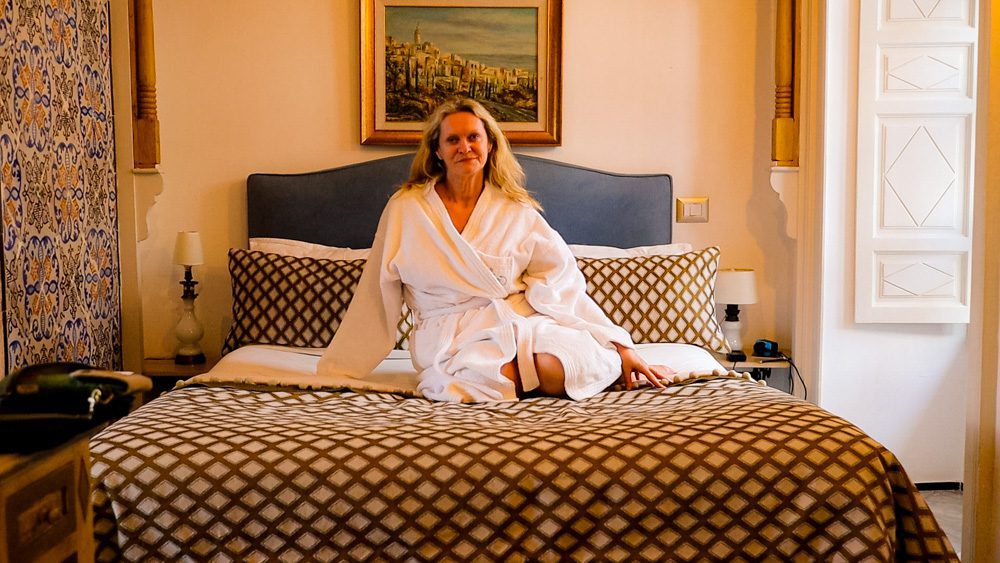 Woman with blonde hair wearing a white dressing gown sitting on a bed with a brown checked cover