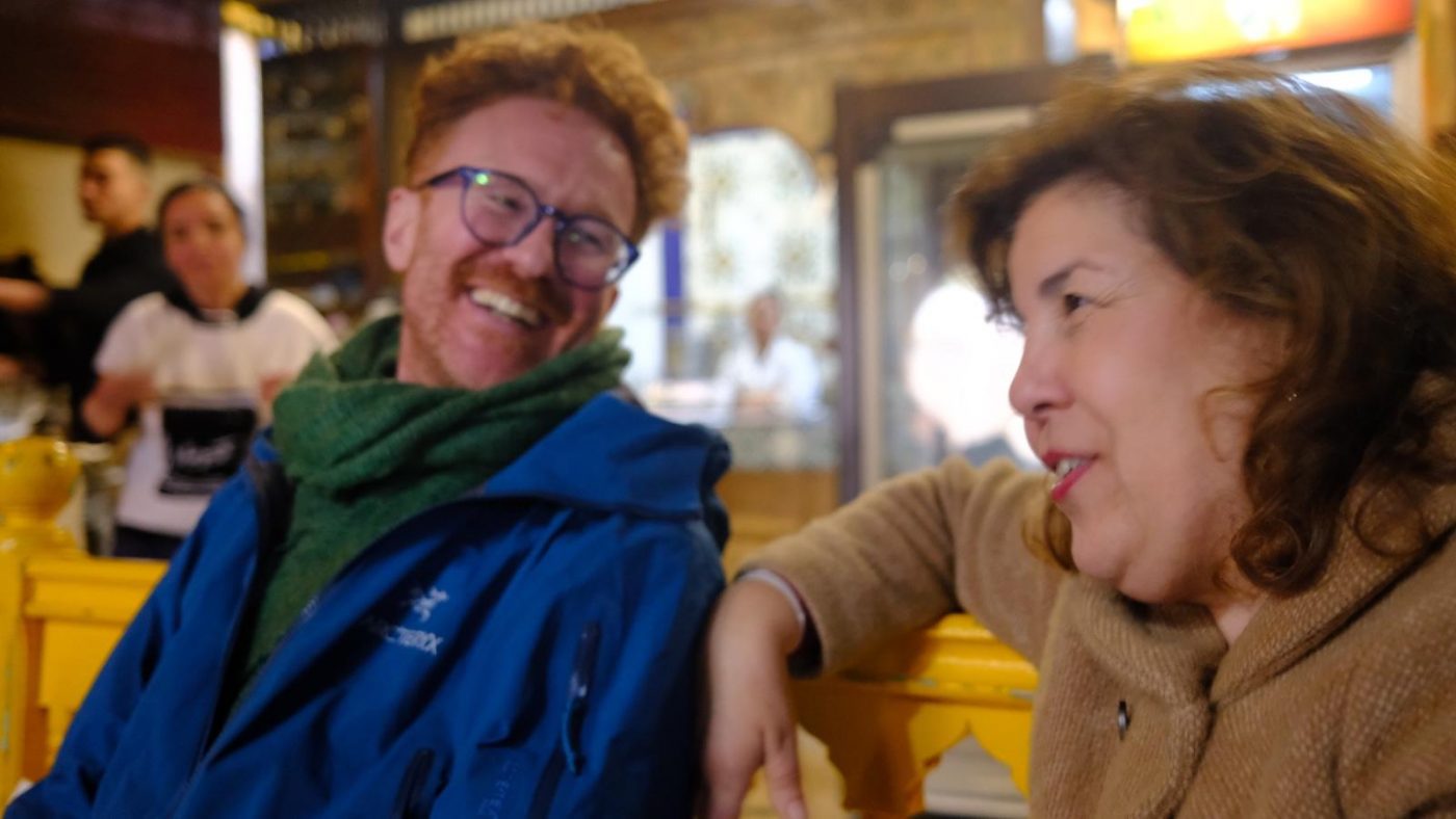 Sawa Taste of Tunisia: Explore the amazing food of Tunisia (2024) 2 Jamie and Lamia, founders of Sawa Taste of Tunisia. Jamie on the left wearing a blue jacket, green scarf and blue glasses. He has ginger hair and a stubble. Lamia, on the right, wears a brown woolen jacket and had red lipstick. They are sitting on a bright yellow bench which just shows behind them