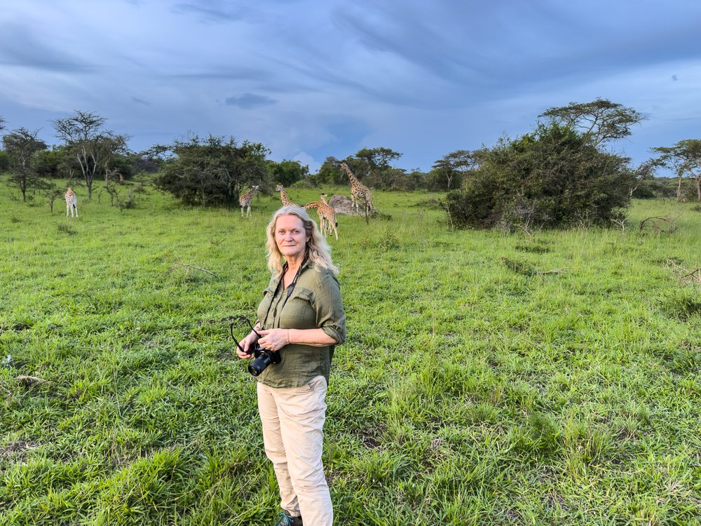 Luxury Stay At Mihingo Lodge, Lake Mburo, Uganda 19 Larch wearing a green shirt and beige trousers and holding a camera with giraffes in the background