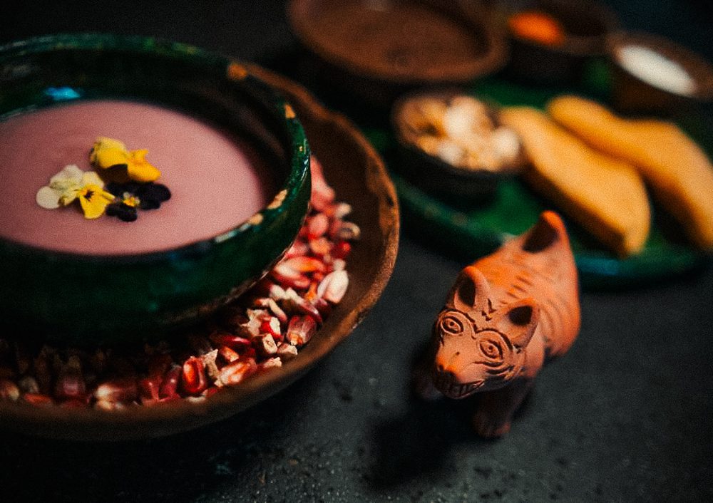 A vibrant, artistic display of food featuring a colorful soup garnished with flowers, surrounded by corn kernels and a small decorative animal figure.